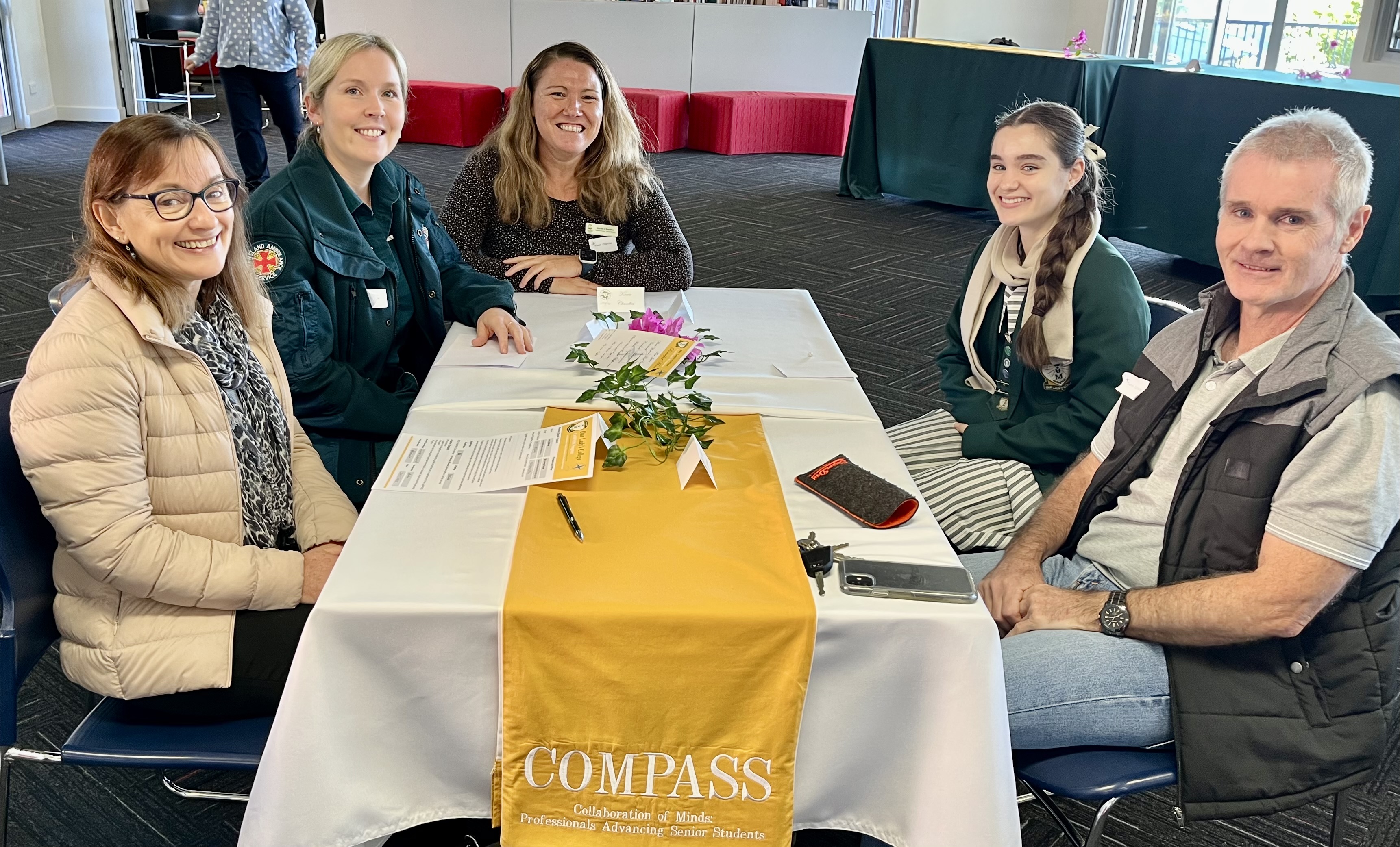 Five people seated around a table at an indoor event, smiling toward the camera. The table holds event materials, a plant centrepiece, and a banner reading “COMPASS – Collaboration of Minds: Professionals Advancing Senior Students”.